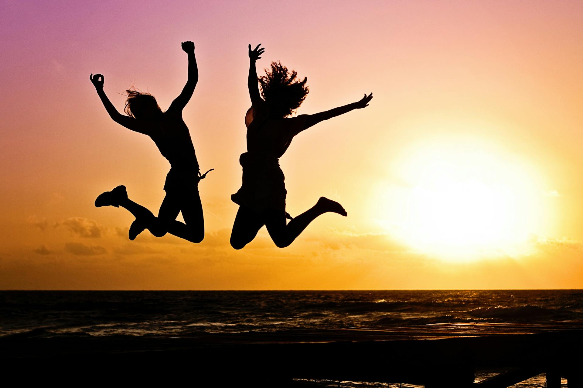 Two people jumping at the beach with the sun setting behind them by Jill Wellington